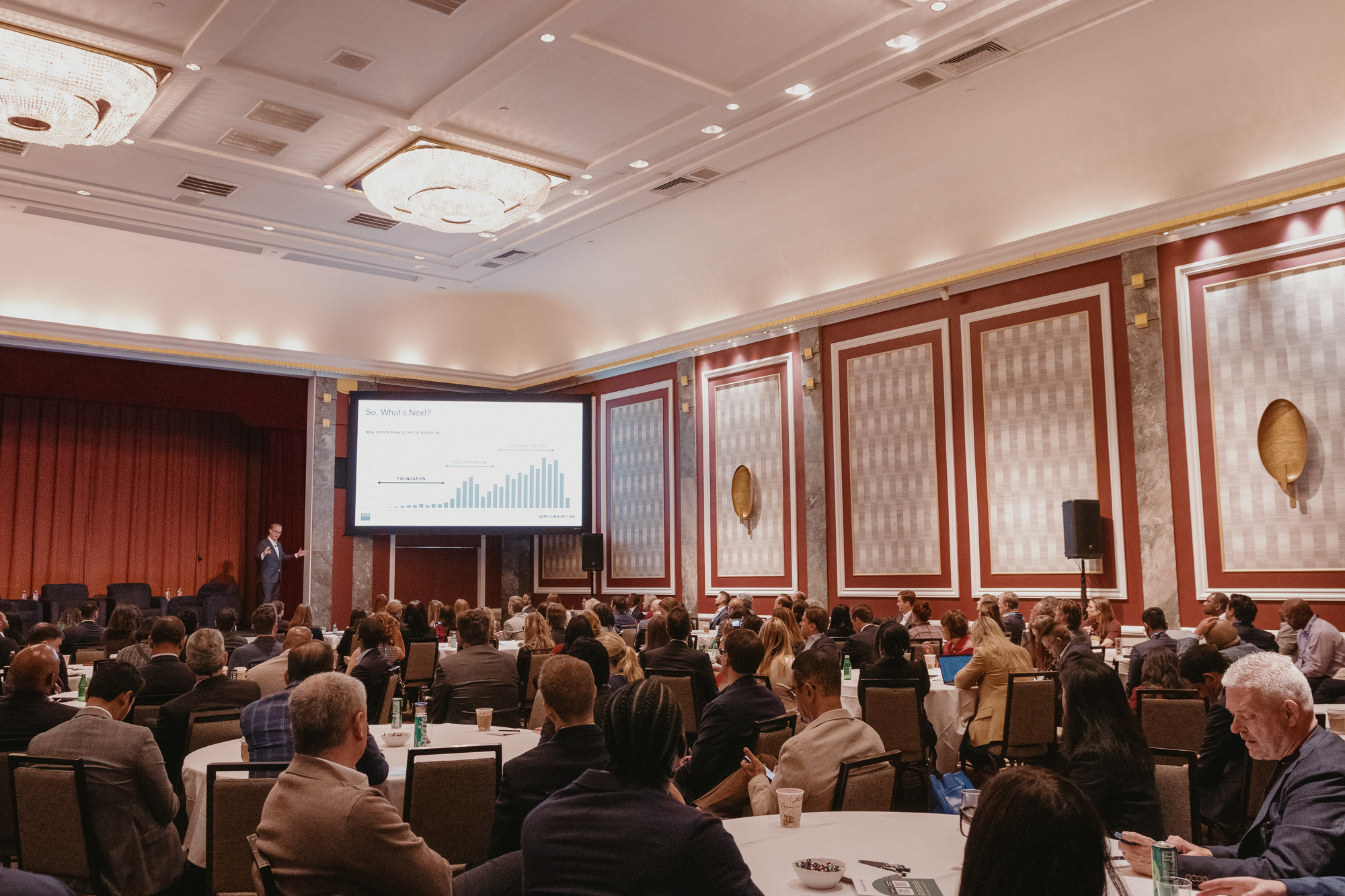 ピーター・ブラフマンがステージで存在している間、丸いテーブルに座っている人々で満たされた大きな会議のボールルーム