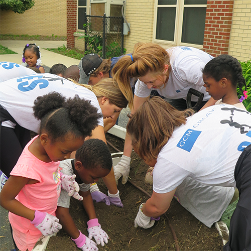 大人と子供のグループは、庭のベッドで一緒に働き、手袋を着用しながら掘り、植えます。大人の多くは、「CGM」と「Chicago Cares」と書かれたロゴのあるマッチングシャツを着ています。