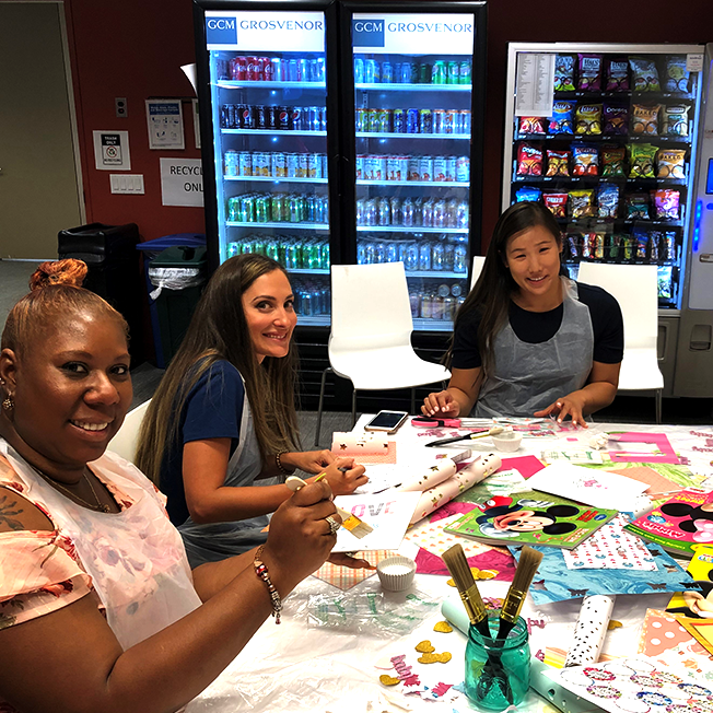 女性のグループは、マーカー、ステッカー、紙など、芸術品や工芸品の備品で覆われたテーブルの周りに座っています。彼らは笑顔で創造的なプロジェクトに取り組んでいます。