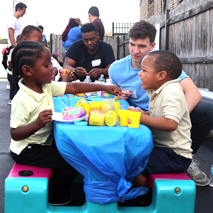 2人の若い男の子が小さな屋外のテーブルに座って、カラフルなプレイドーと遊んでいる間、2人のボランティアが彼らと交流し、笑顔で活動に従事します。