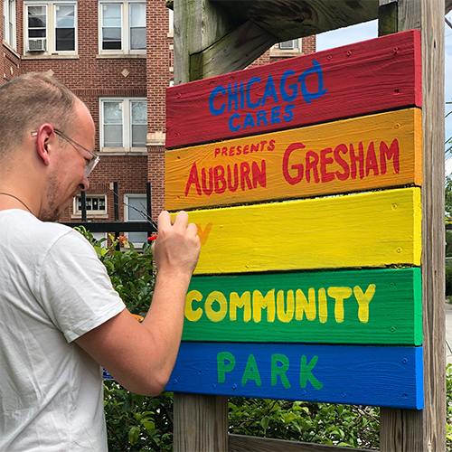 男が明るく大胆な文字で「シカゴ・ケアーズ・プレゼントのオーバーン・グレシャム・コミュニティパーク」と書かれたカラフルな木製の看板を描いています。シーンは住宅の近くの屋外にあります。