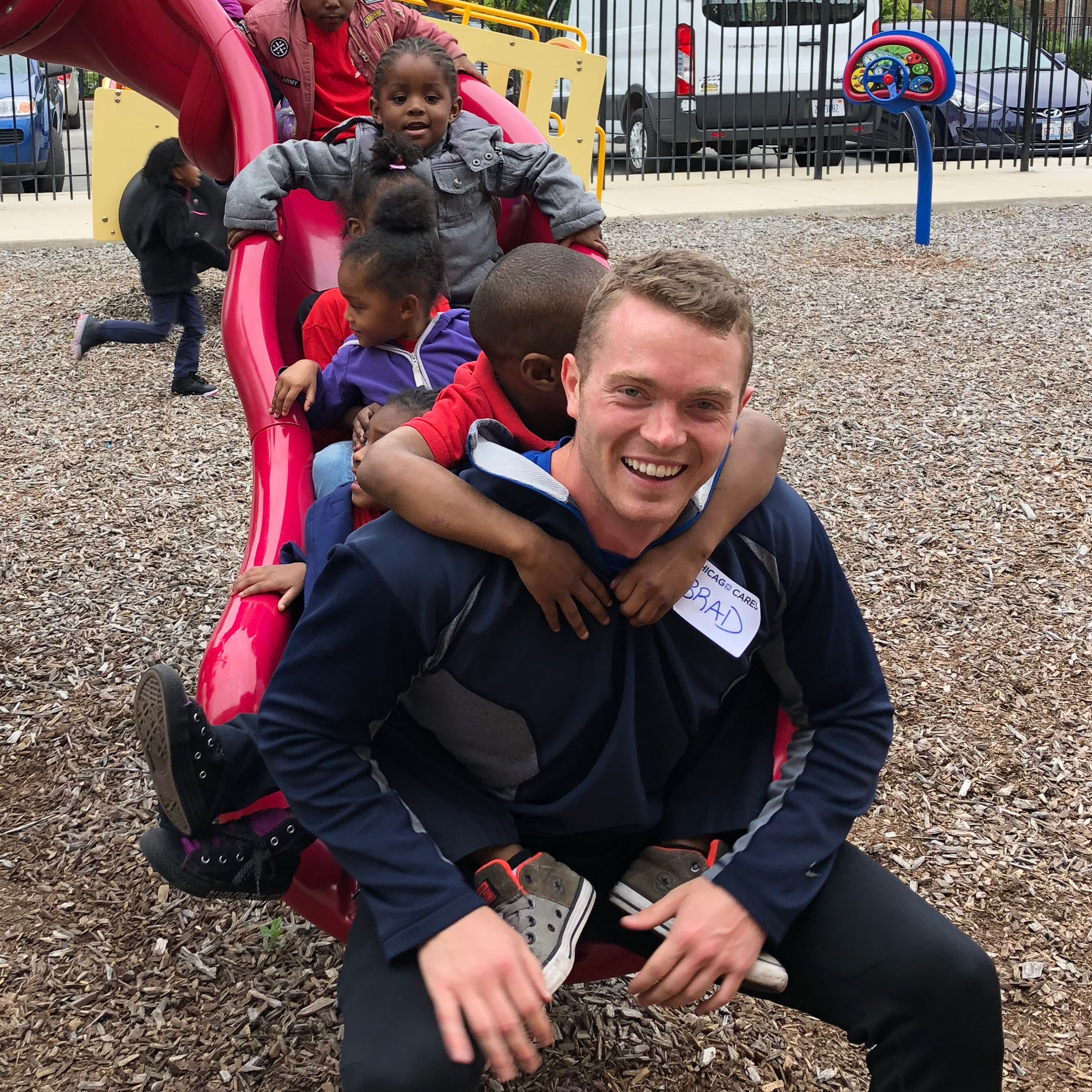 ボランティアが遊び場のスライドの底に座って、子供たちが背中に登って滑り落ち、背景で喜んで遊んでいます。