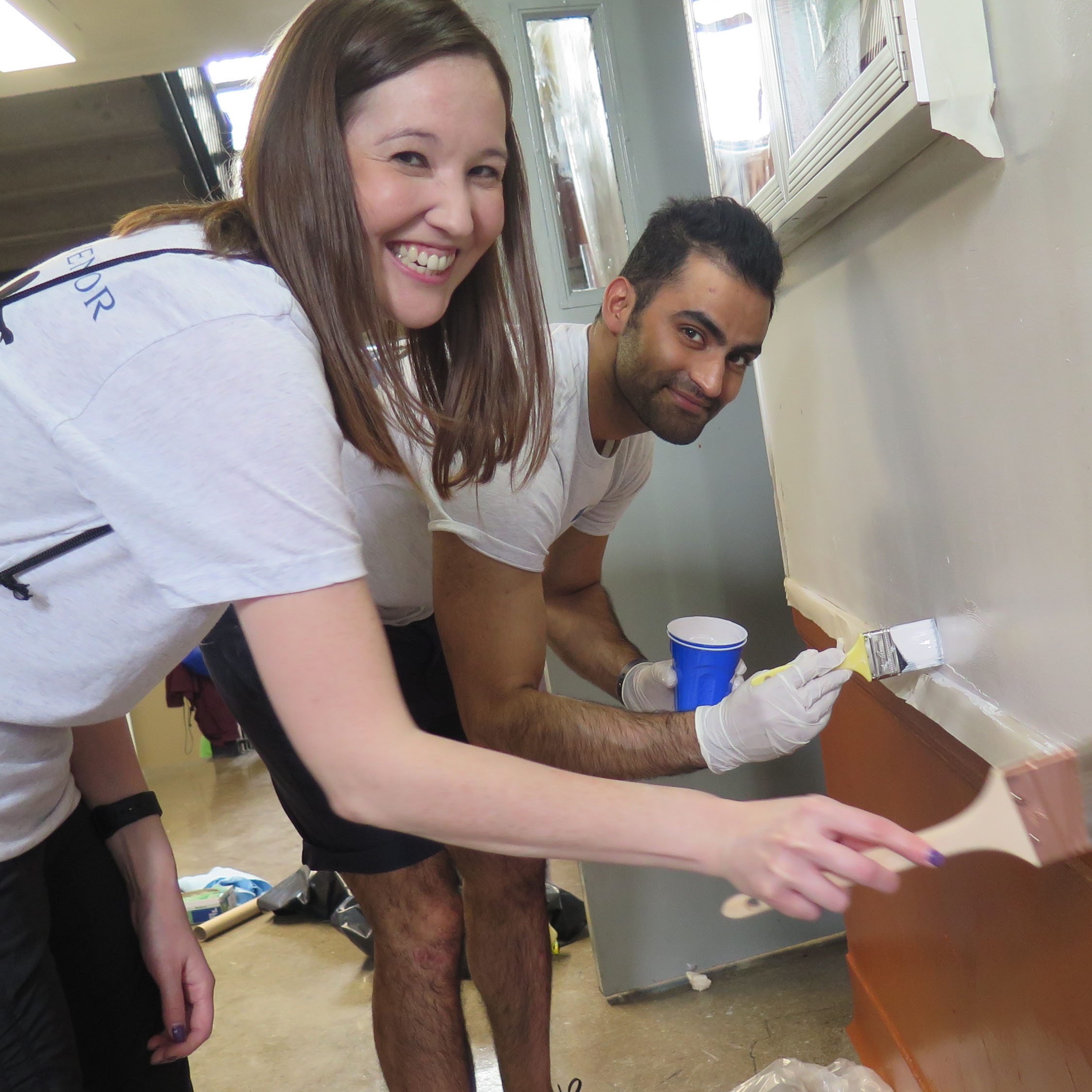 屋内で壁を描くと女性と男性が微笑む。どちらも手袋と白いボランティアのシャツを着ており、陽気でタスクに焦点を合わせているように見えます。